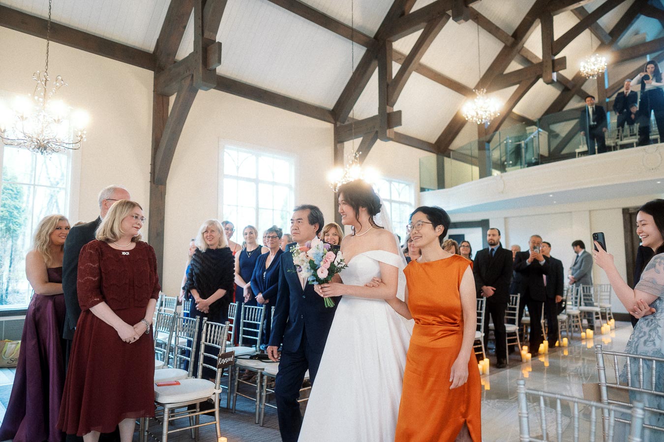 Bride walks down the aisle with two companions during an indoor wedding ceremony.
