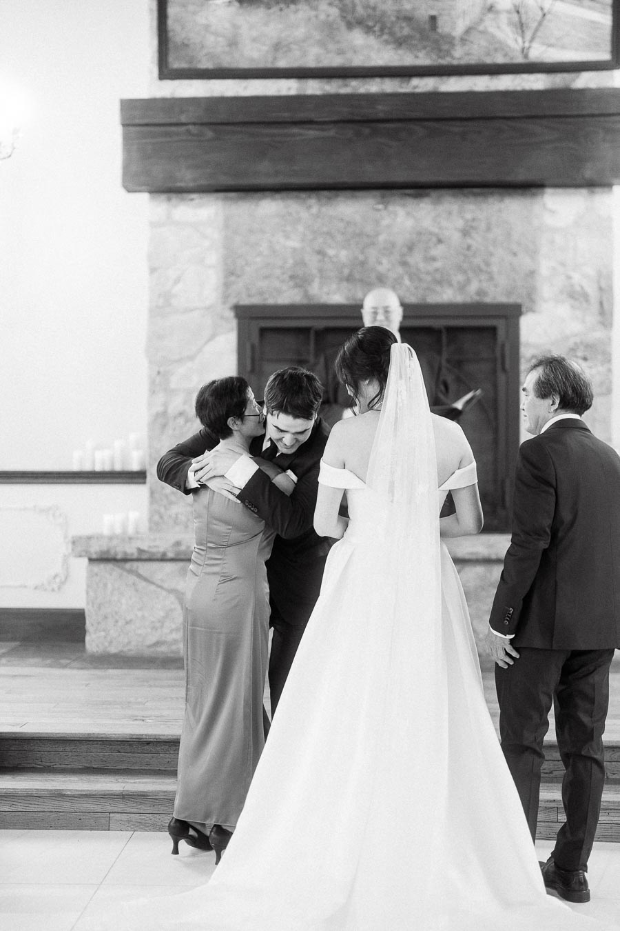 Bride stands nearby as groom embraces a woman in front of a stone fireplace.
