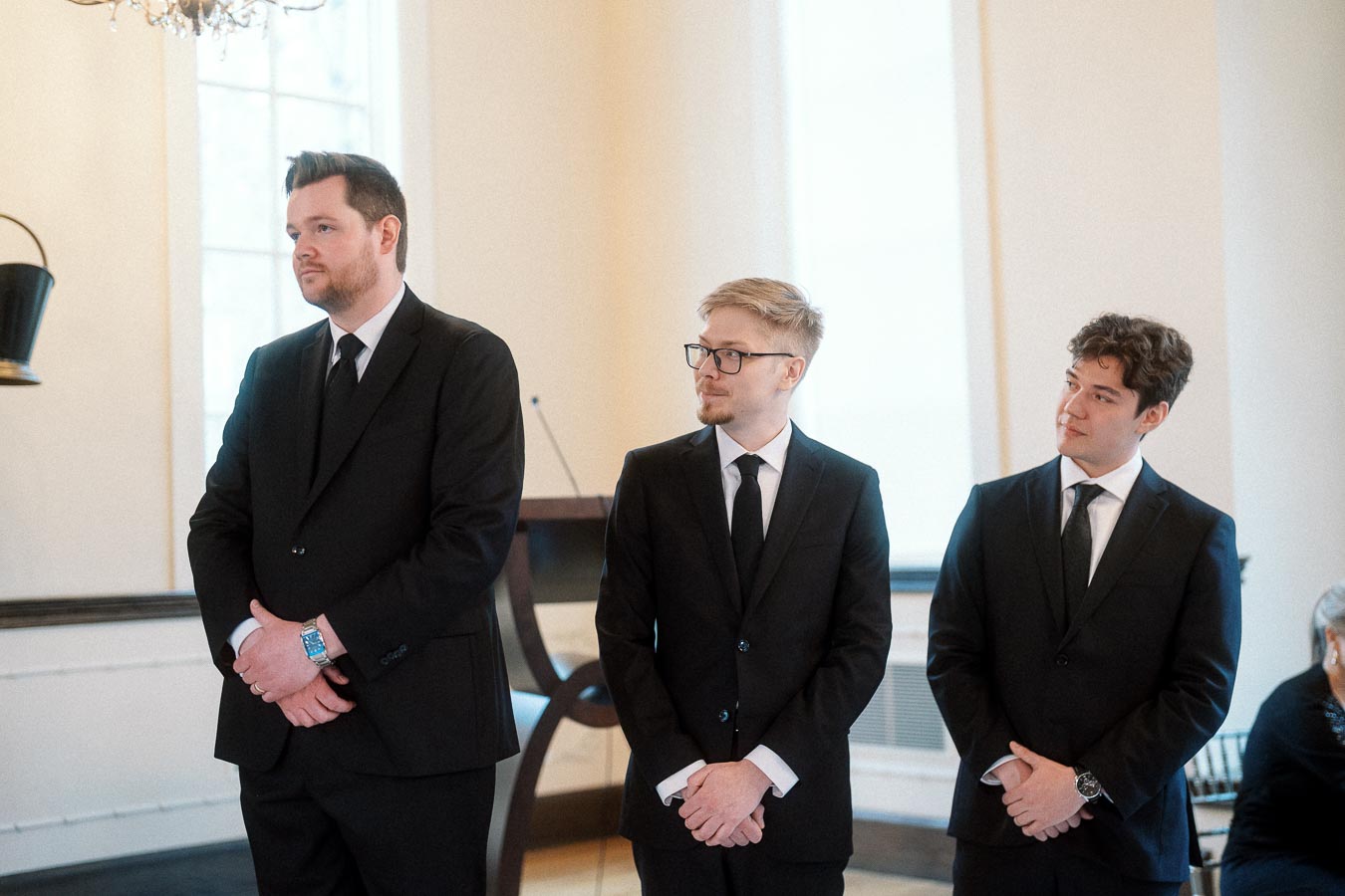 Three men in black suits stand indoors during a formal engagement event.