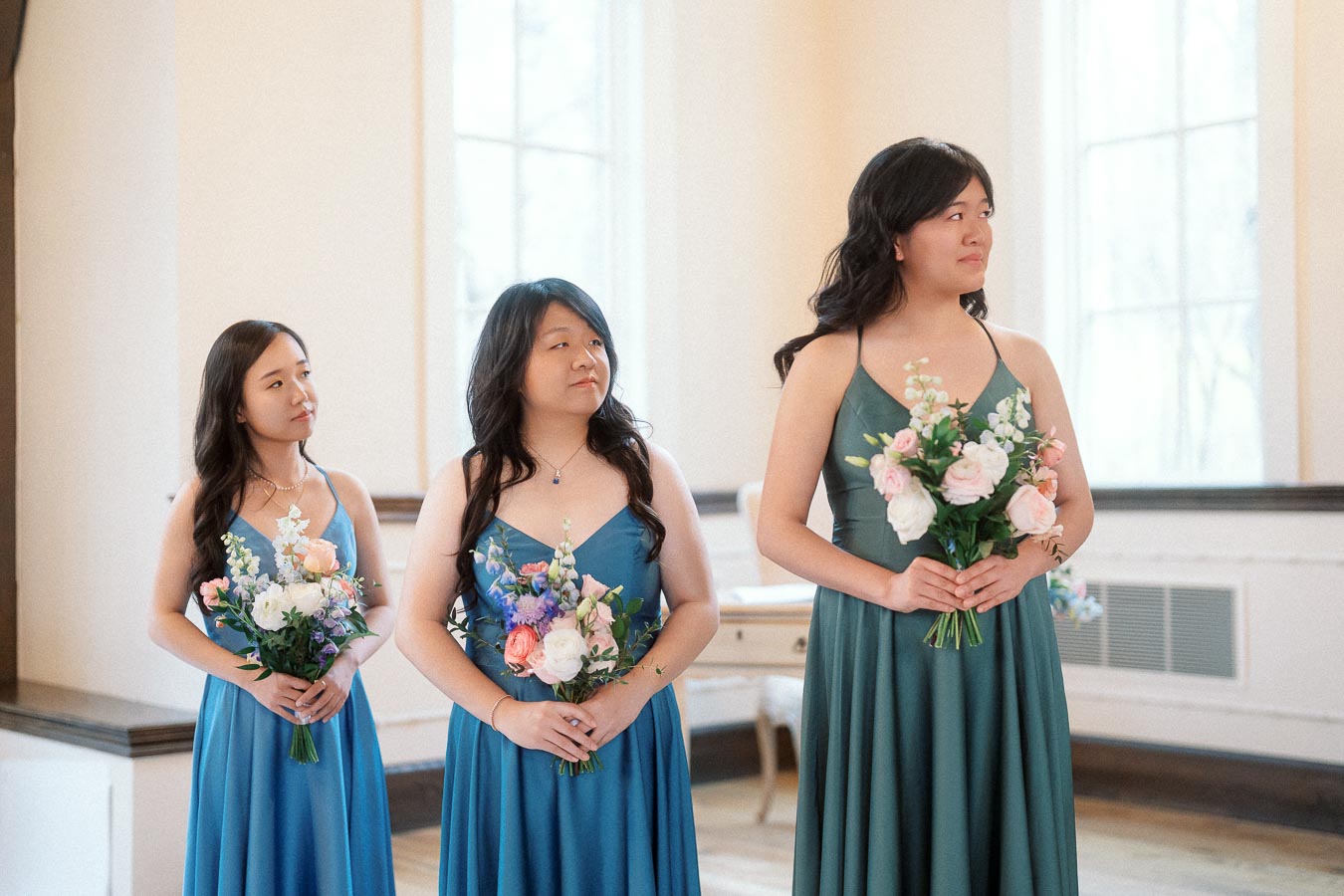 Three women in teal and blue dresses holding floral bouquets indoors.