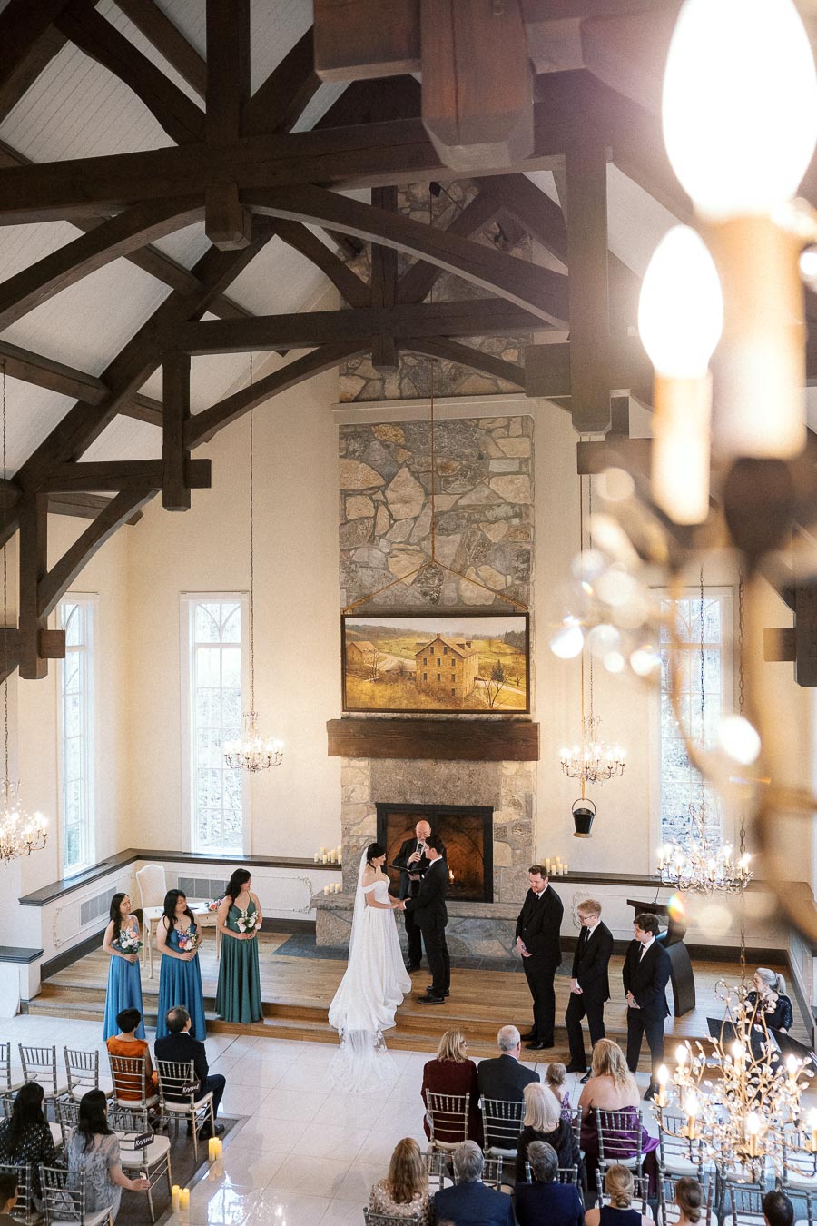 Couple exchanging vows during an indoor wedding ceremony beneath wooden beams and chandeliers.