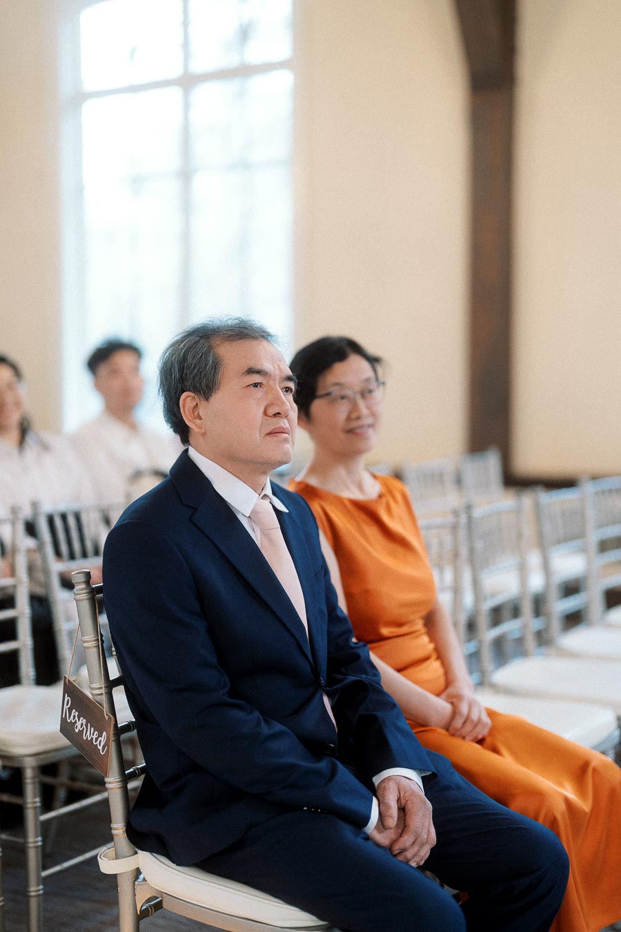 Well-dressed guests seated on reserved chairs during an indoor ceremony.