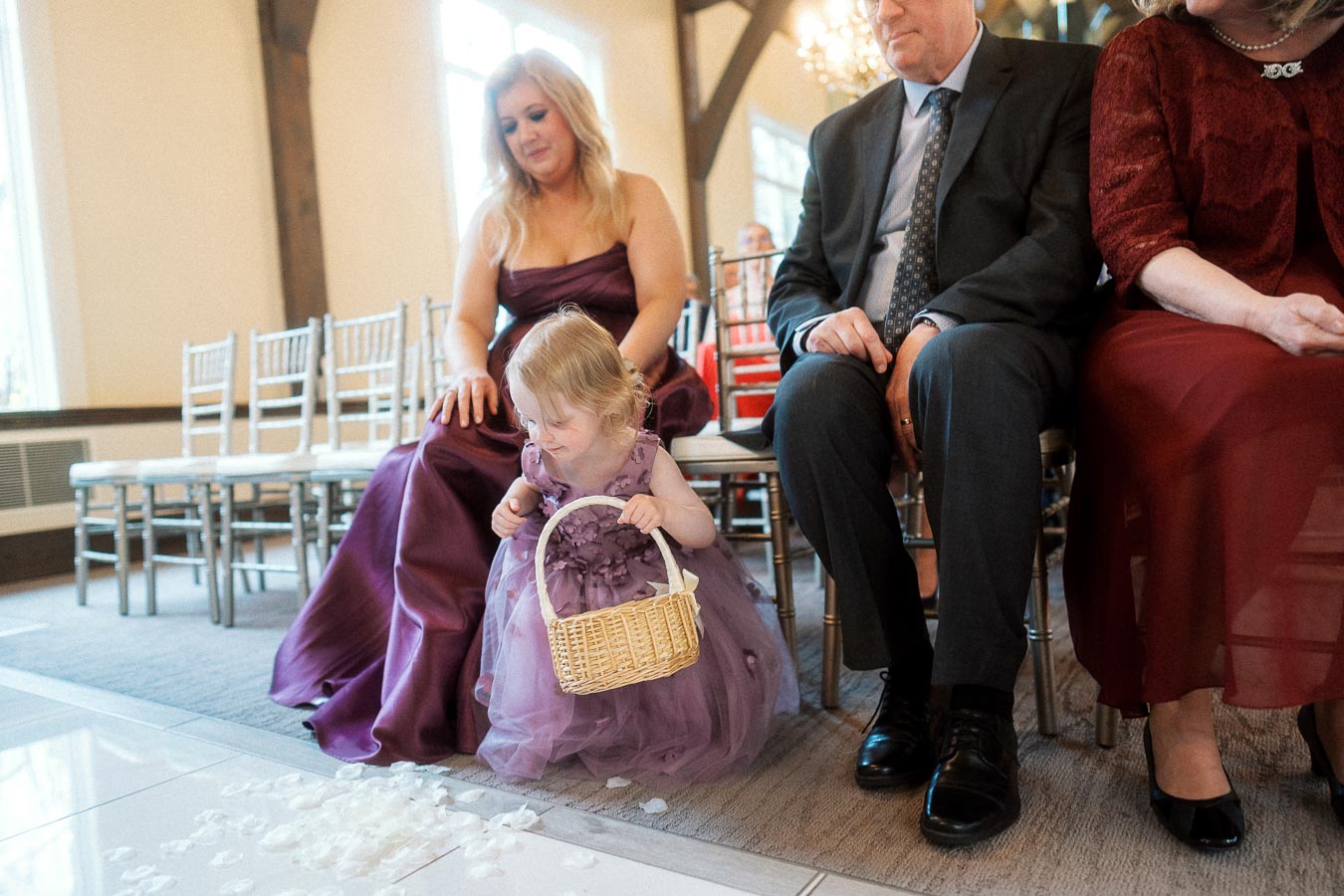 Young flower girl in a purple dress holding a basket beside seated wedding guests