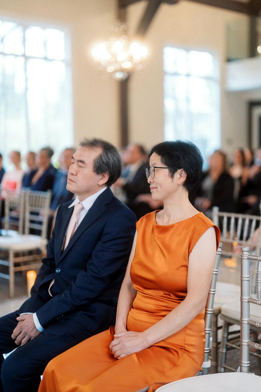 Two seated guests in formal attire during an indoor ceremony