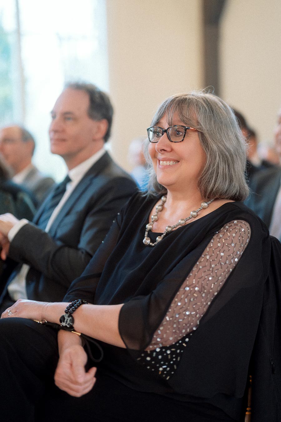 Smiling woman in glasses seated among guests at an indoor event