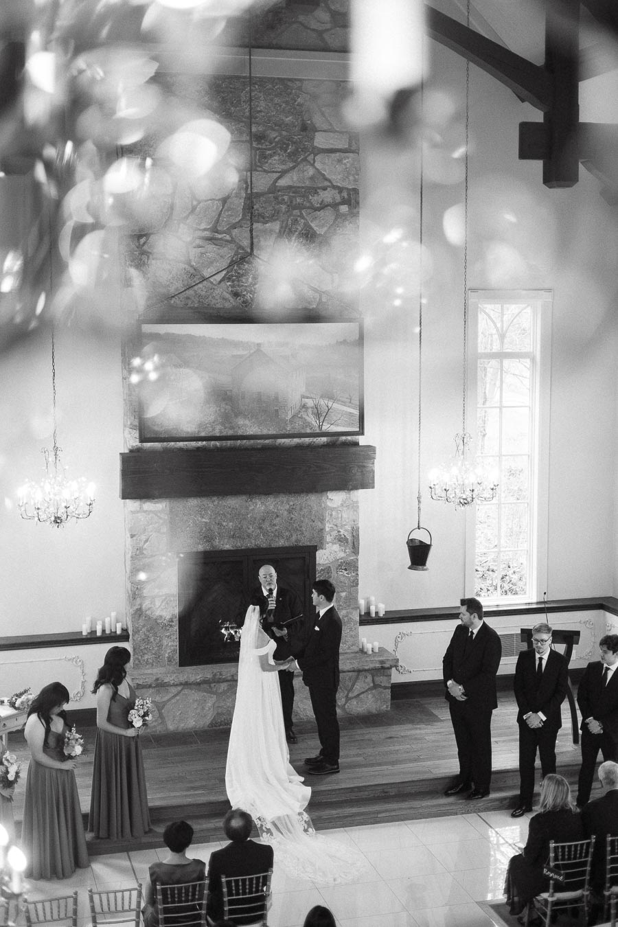 Black-and-white ceremony scene with a couple exchanging vows before a stone fireplace.