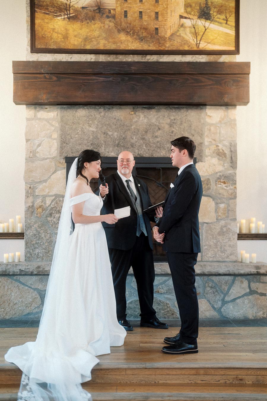 Bride reads vows to groom during an indoor wedding ceremony by a stone fireplace.