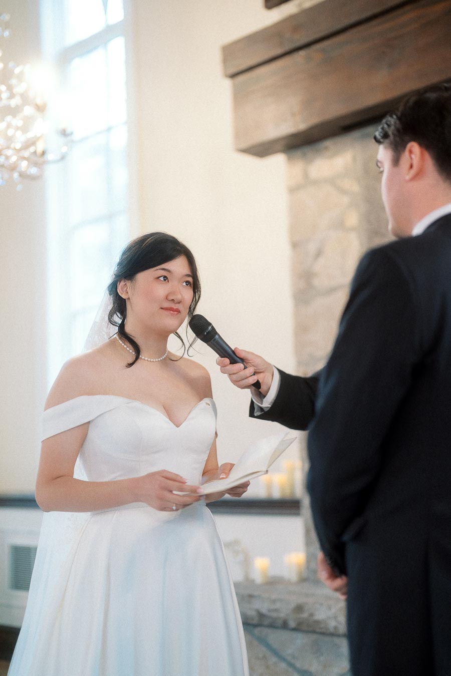 Bride in white dress speaks into a microphone during an indoor ceremony.