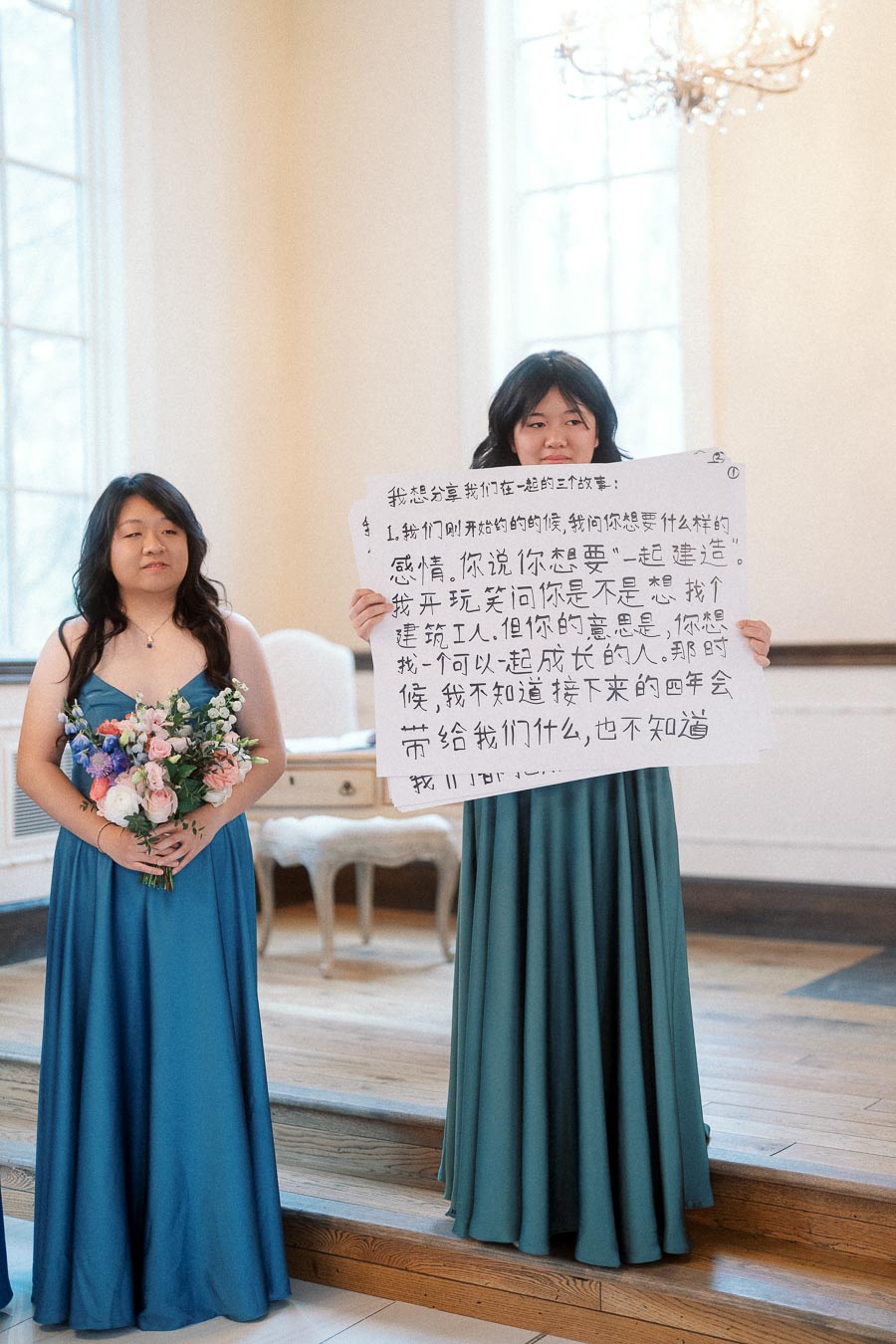 Two women in teal dresses, one holding flowers and one holding a handwritten sign.