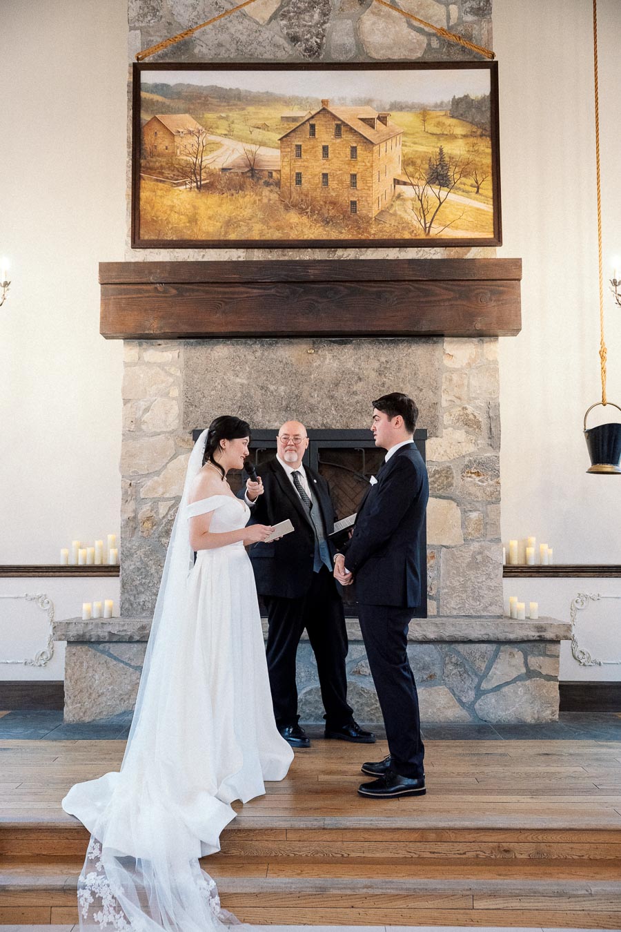 Couple in wedding attire exchanging vows before an officiant by a stone fireplace