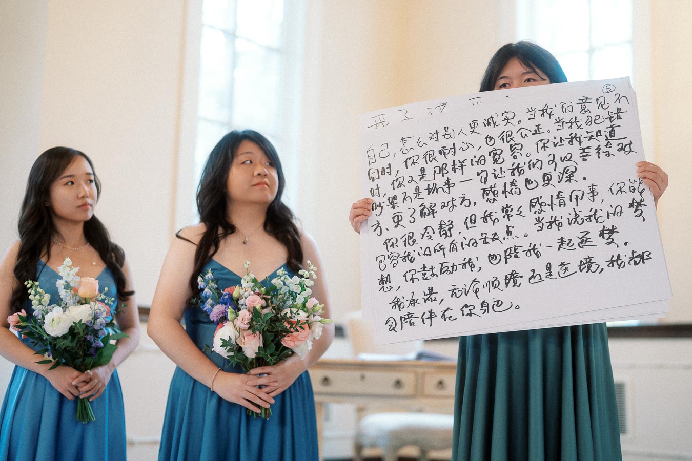 Bridesmaids in blue dresses hold bouquets while one displays a handwritten sign.
