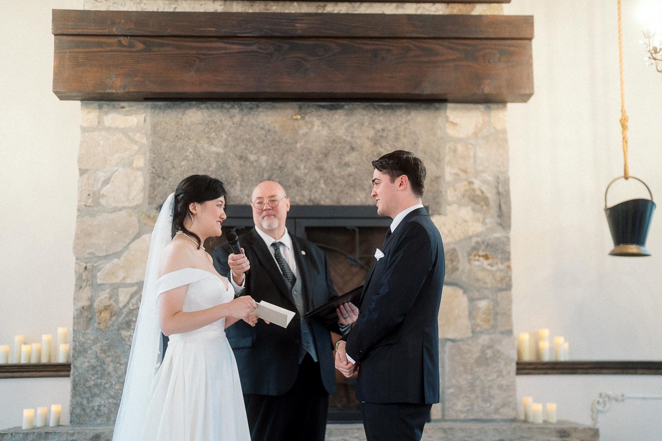 Bride reads vows to groom during an indoor ceremony by a stone fireplace.