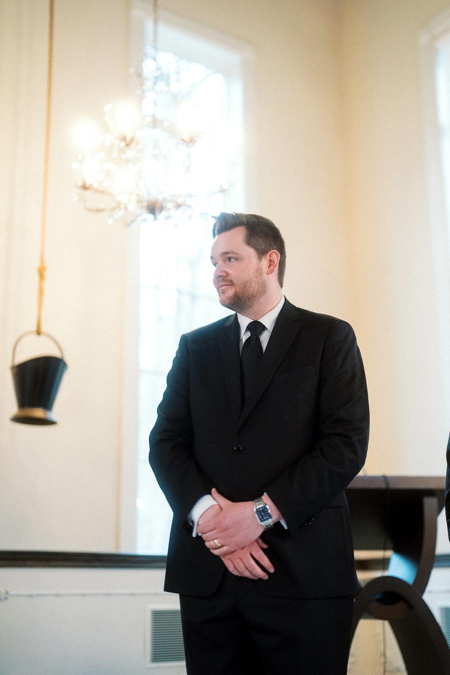 Man in a black suit standing indoors near tall windows and a chandelier