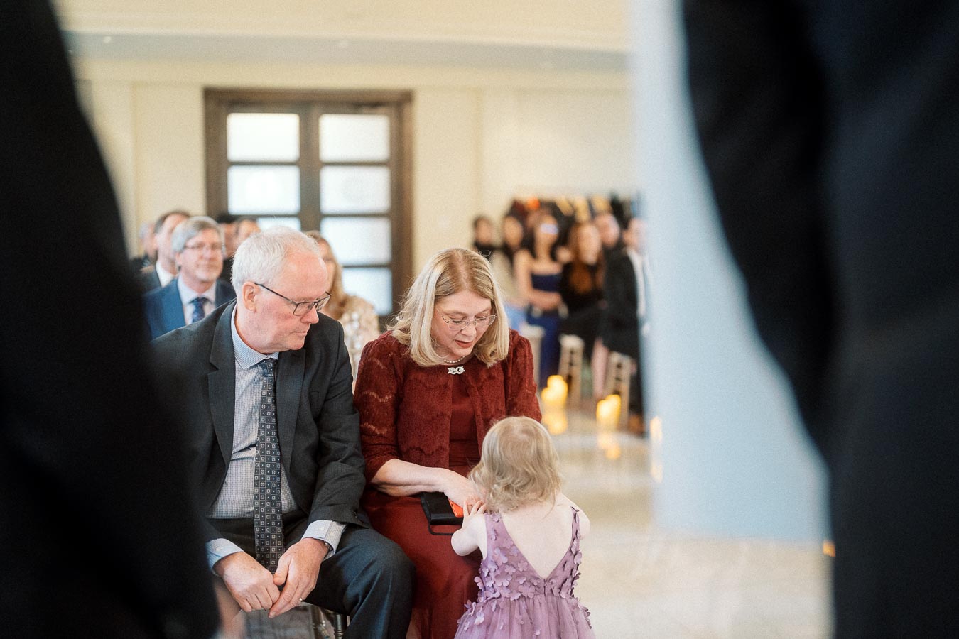 Child in a purple dress standing with seated guests during an indoor ceremony