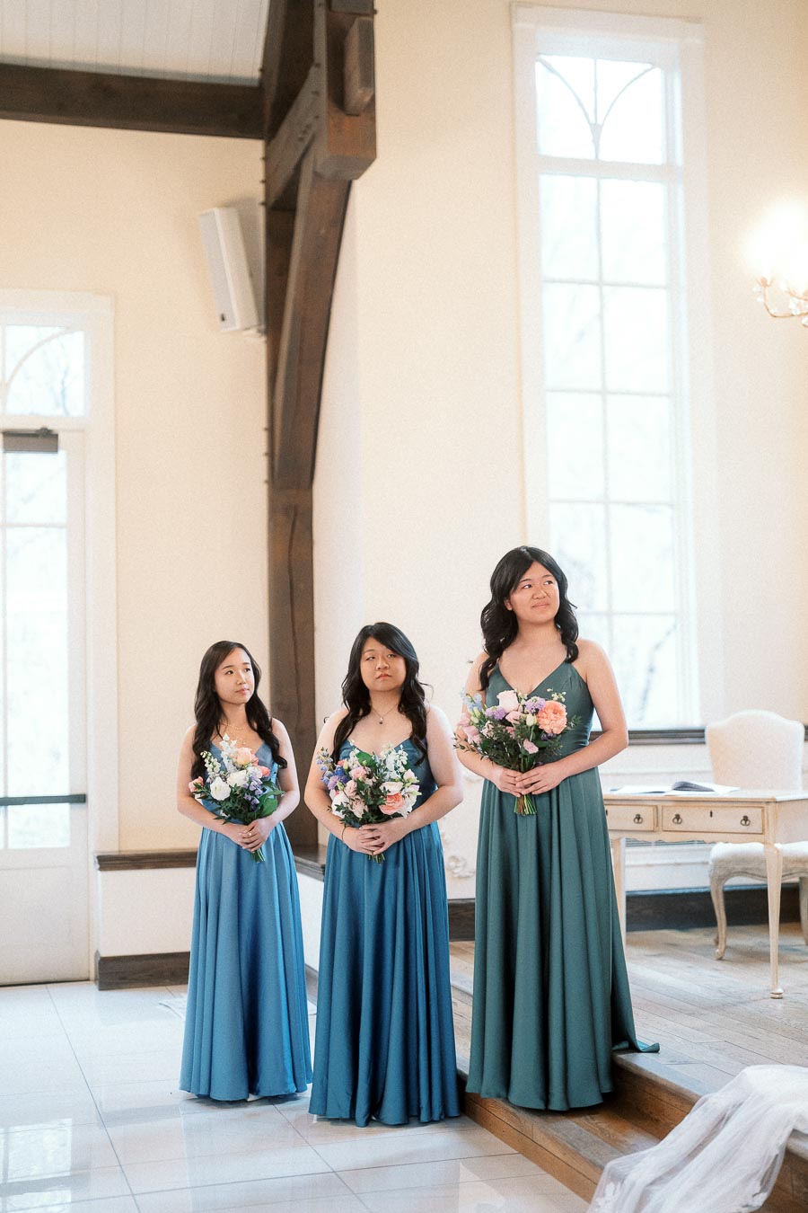 Three bridesmaids in teal dresses holding bouquets inside a bright room