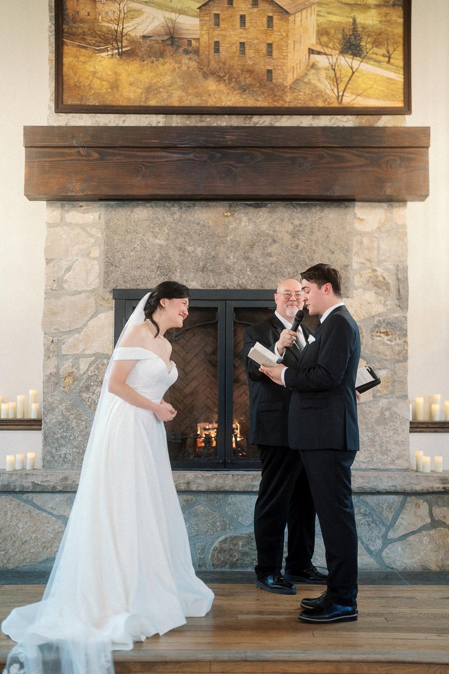 Bride laughs as groom reads vows beside an officiant and stone fireplace.