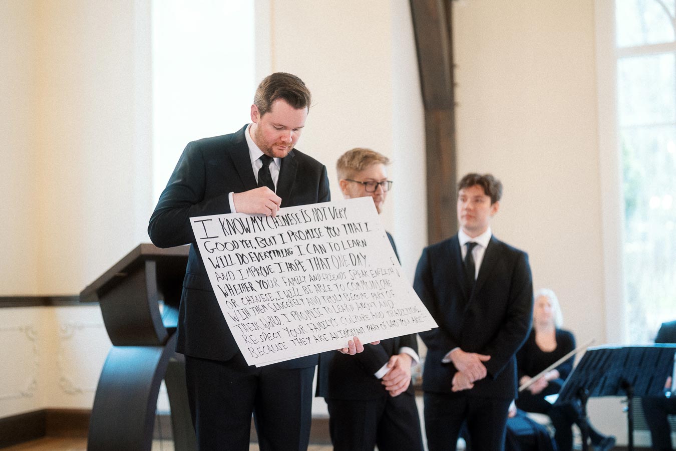 Man in a black suit reading from a handwritten sign during an indoor ceremony