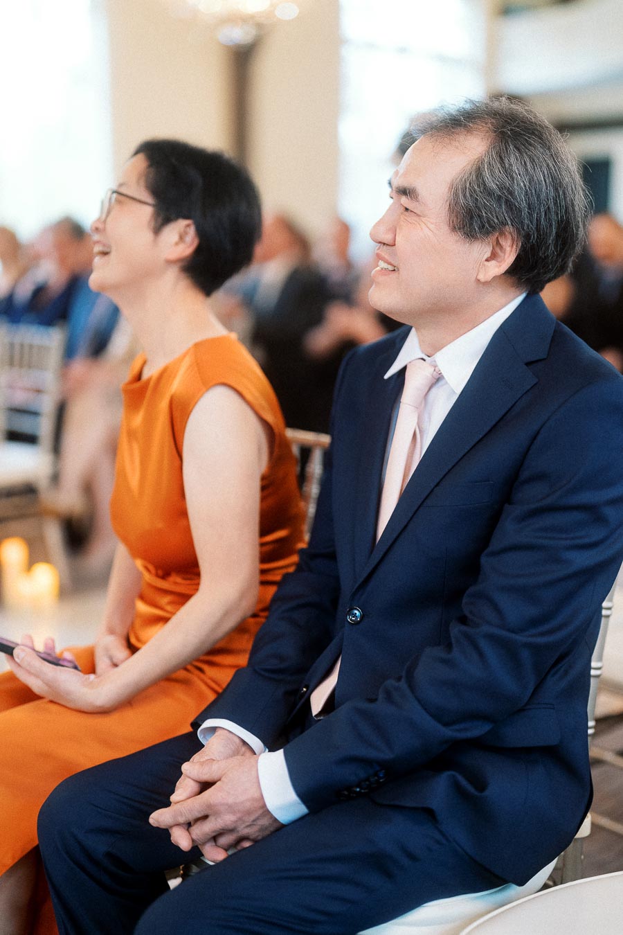 Two formally dressed guests smiling while seated at an indoor ceremony