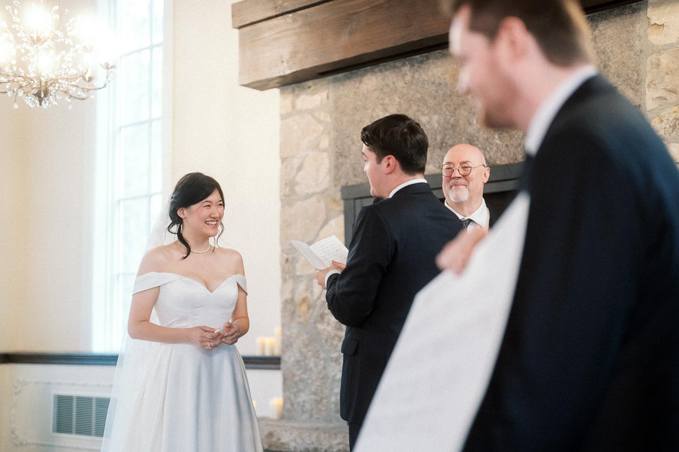 Bride smiling during an indoor wedding ceremony beside a stone fireplace.