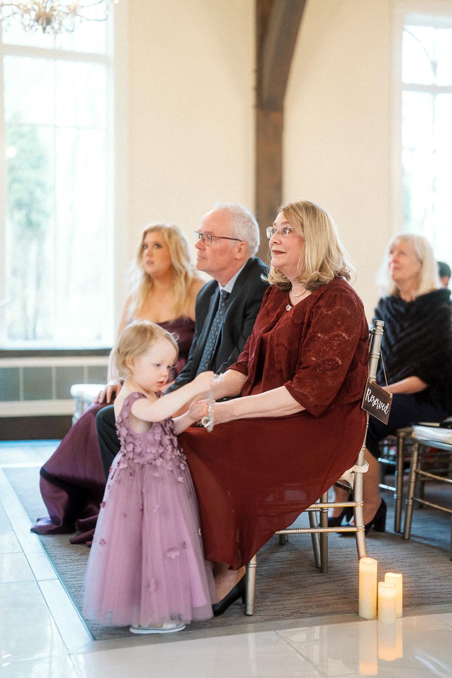 Young flower girl stands beside seated guests during an indoor ceremony.