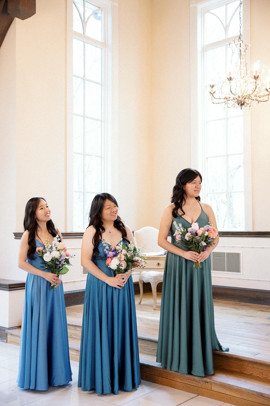 Three women in long dresses holding bouquets in a bright room with tall windows