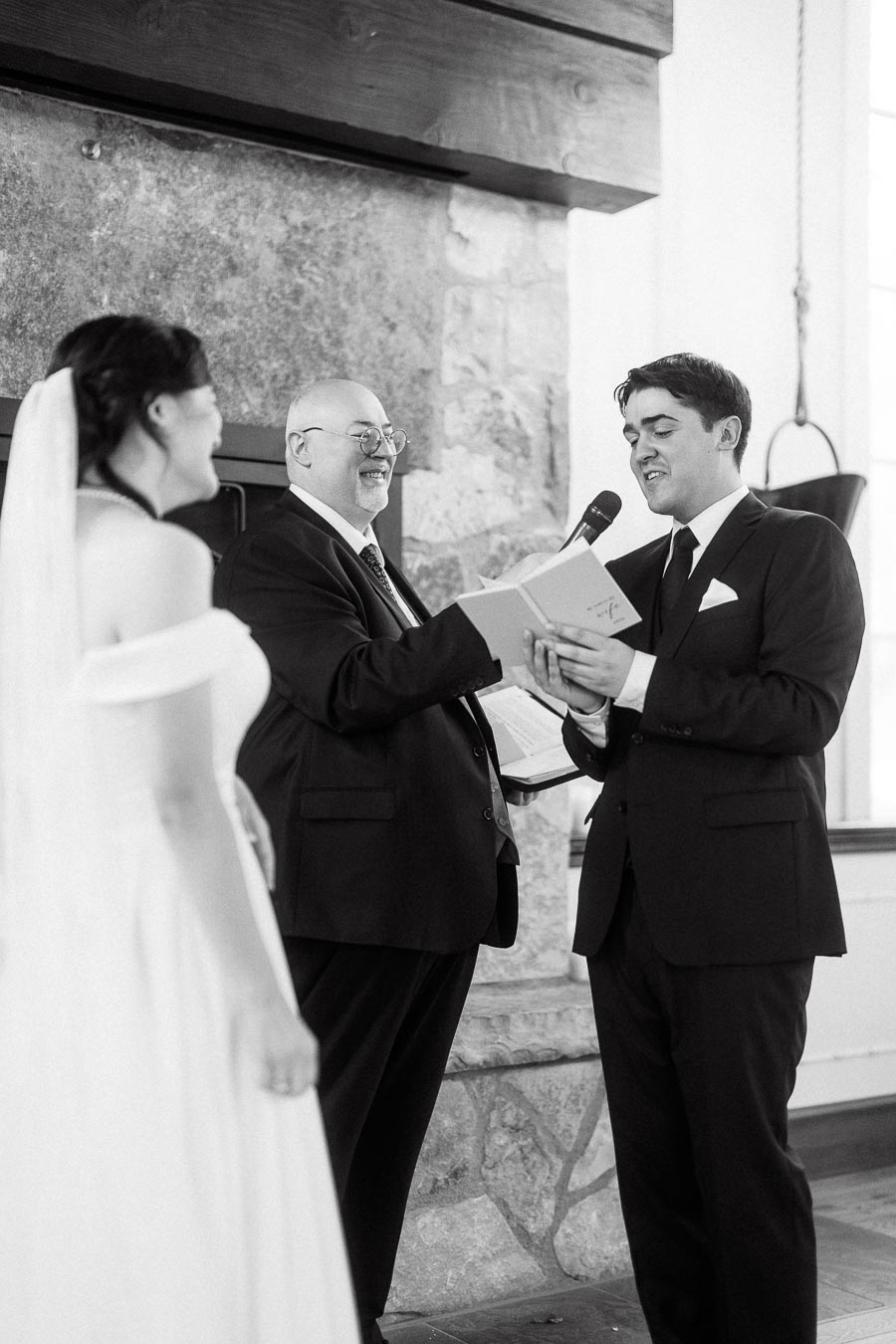Groom reads from a booklet beside an officiant as the bride smiles during the ceremony.