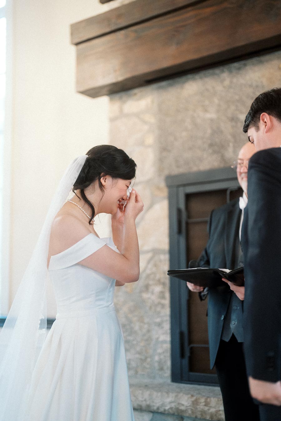 Bride wiping away tears during an indoor ceremony beside the groom and officiant
