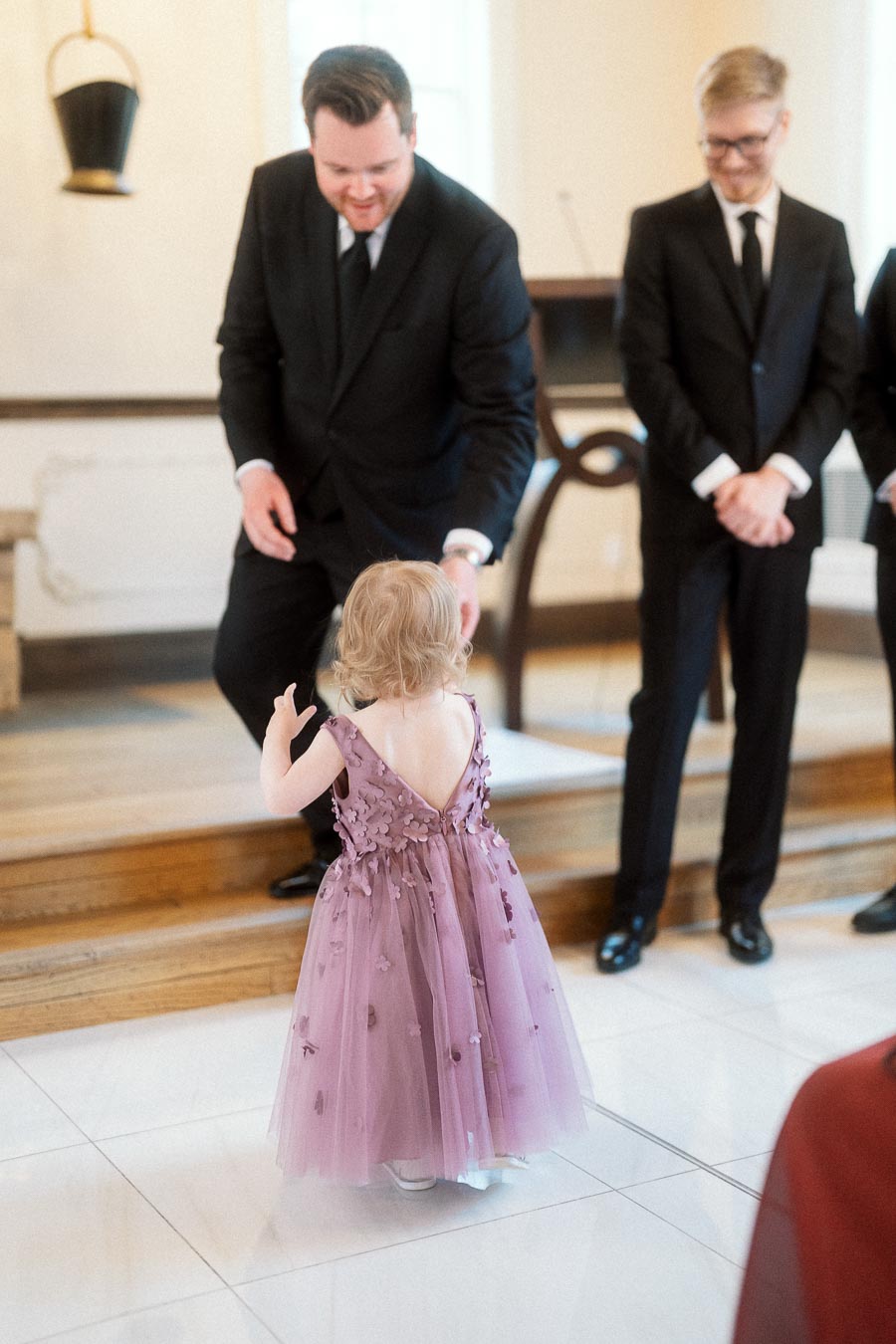 Young child in a lavender dress walking toward a man in a black suit indoors