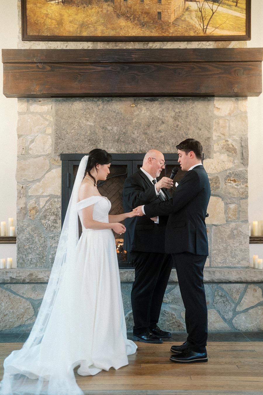 Couple exchanging rings during an indoor wedding ceremony by a stone fireplace