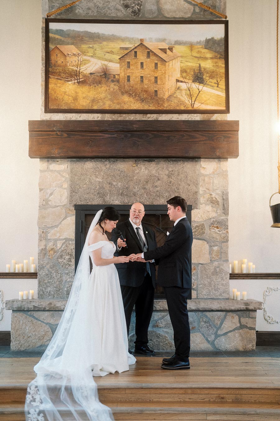 Bride and groom exchange rings during an indoor wedding ceremony by a stone fireplace.