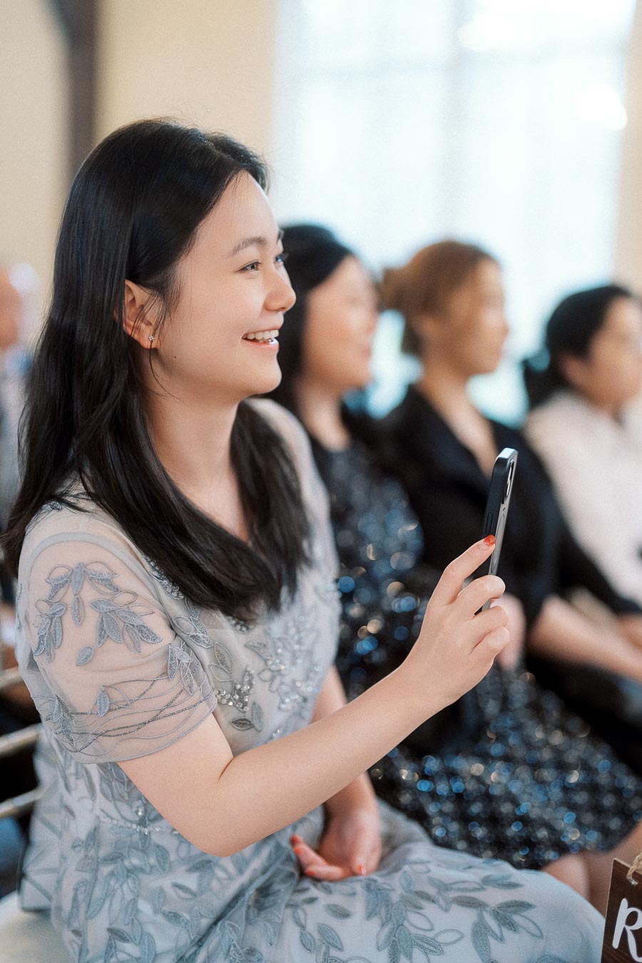 Smiling woman seated indoors holding a smartphone among other guests.