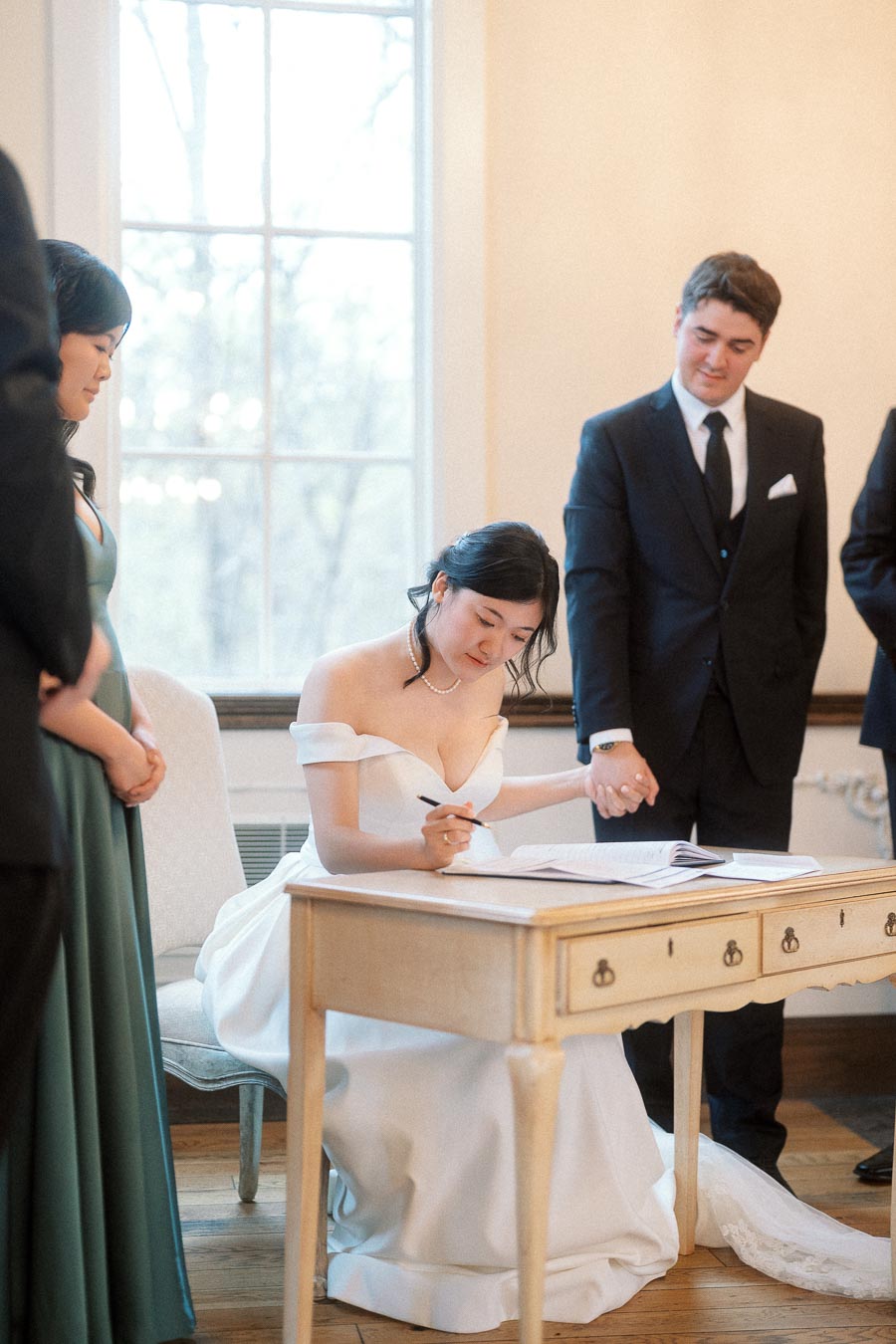 Bride signs a document while holding hands with groom during an indoor ceremony.