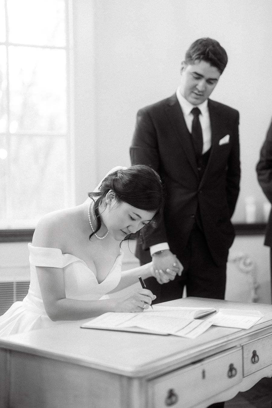 Bride signs a document while holding hands with groom in a bright room