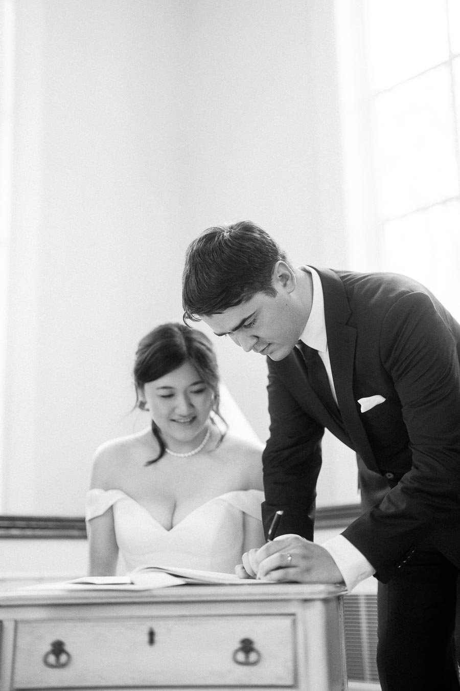 Couple in formal attire signing a book beside a bright window.