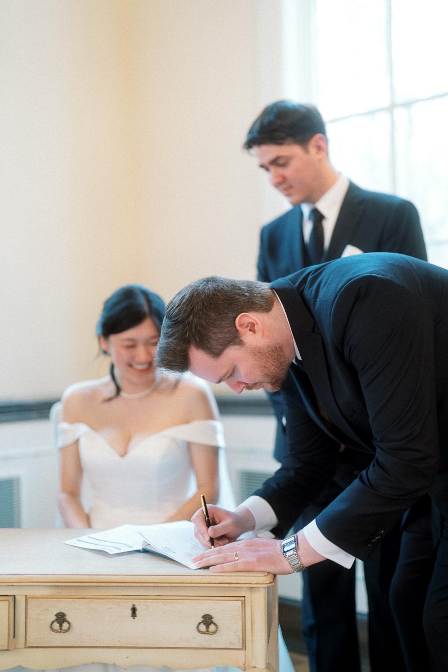 Man in a dark suit signs a document beside a seated woman in a white dress.