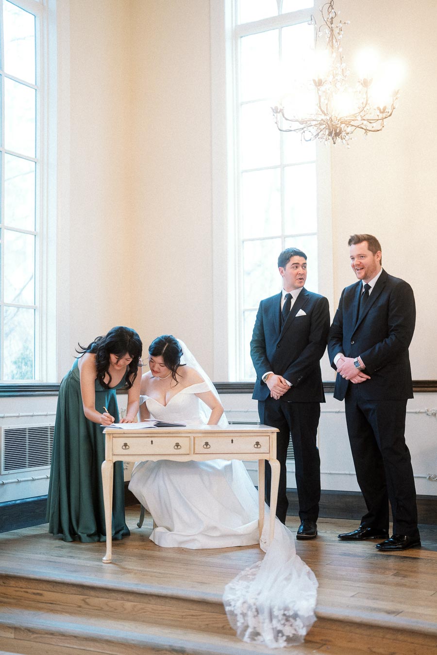 Bride signs documents at a small desk while attendants stand nearby.