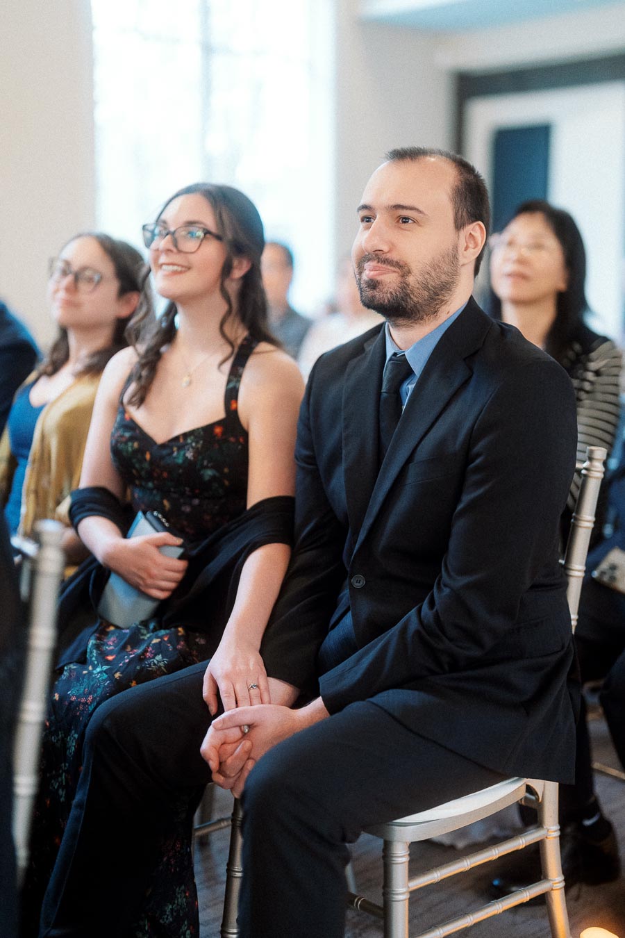 Couple seated together at an indoor engagement event, holding hands among guests.