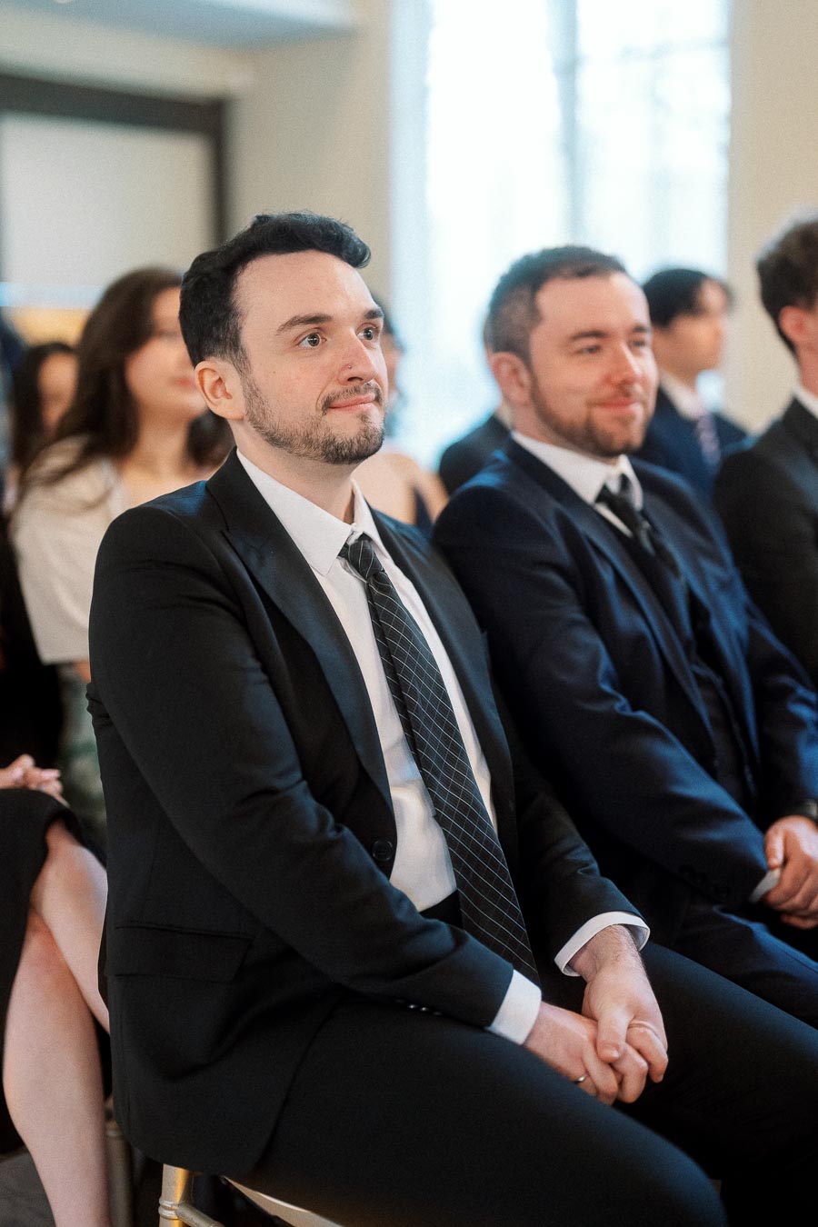 Man in a black suit seated among guests at a formal indoor ceremony