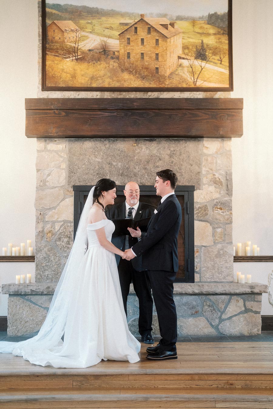 Bride and groom holding hands during an indoor wedding ceremony by a stone fireplace