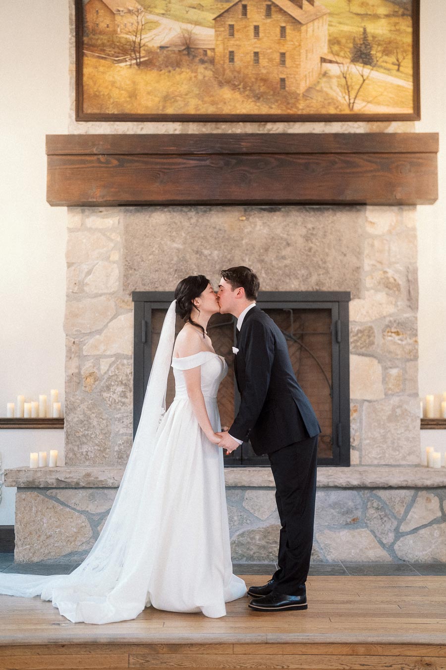 Couple in wedding attire kissing while holding hands in front of a stone fireplace