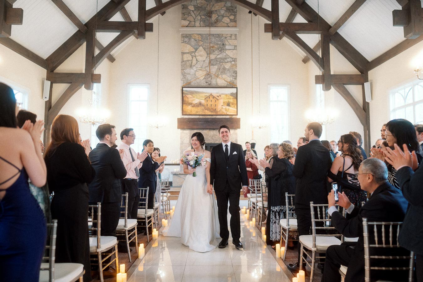 Bride and groom walk down the aisle as guests applaud inside a chapel.