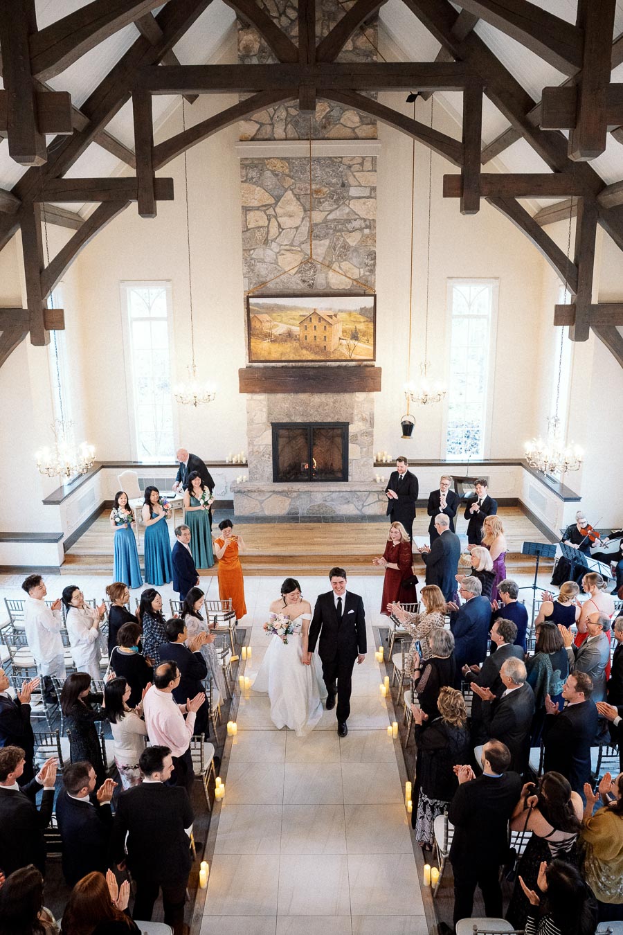 Newly married couple walking down the aisle as guests applaud in a timber-framed ceremony hall