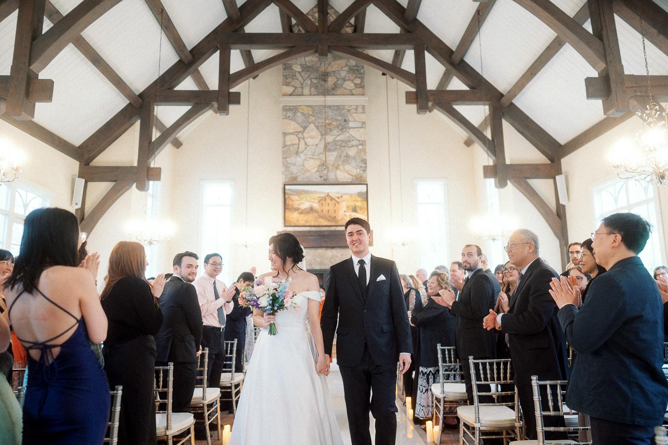 Newlyweds walk down the aisle as guests applaud inside a timber-beamed chapel.