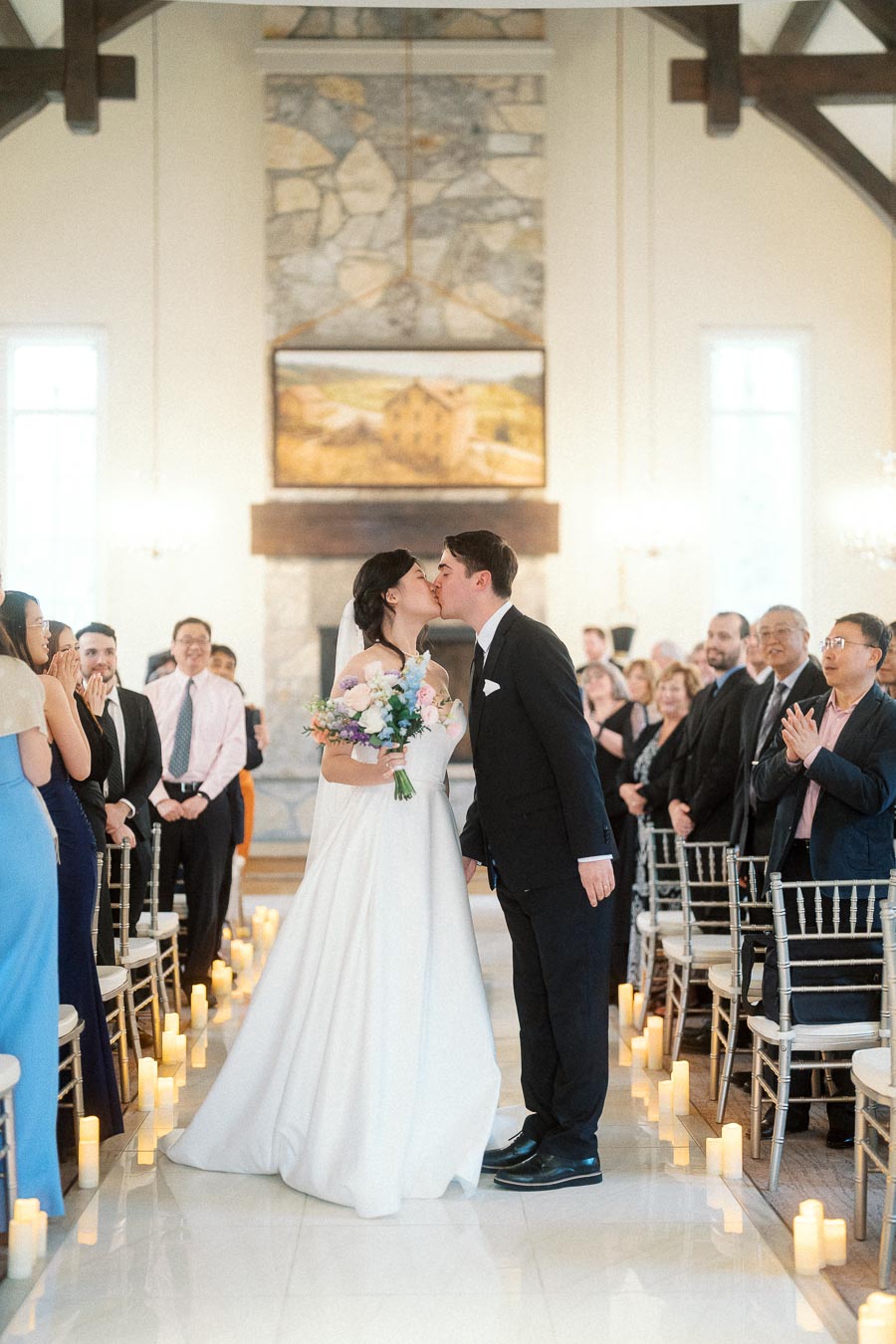 Bride and groom kiss in a candlelit aisle while guests applaud.