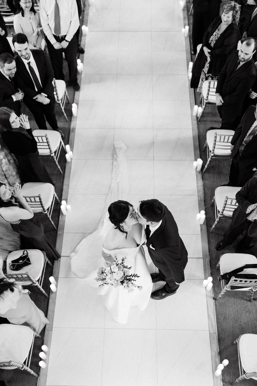 Bride and groom kiss in a candle-lined aisle surrounded by seated guests.