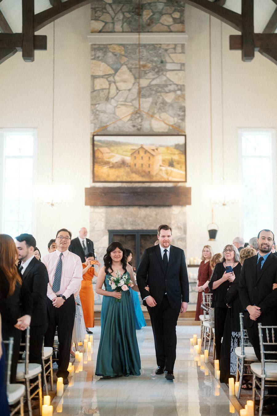 Wedding attendants walk down a candle-lined aisle inside a chapel.