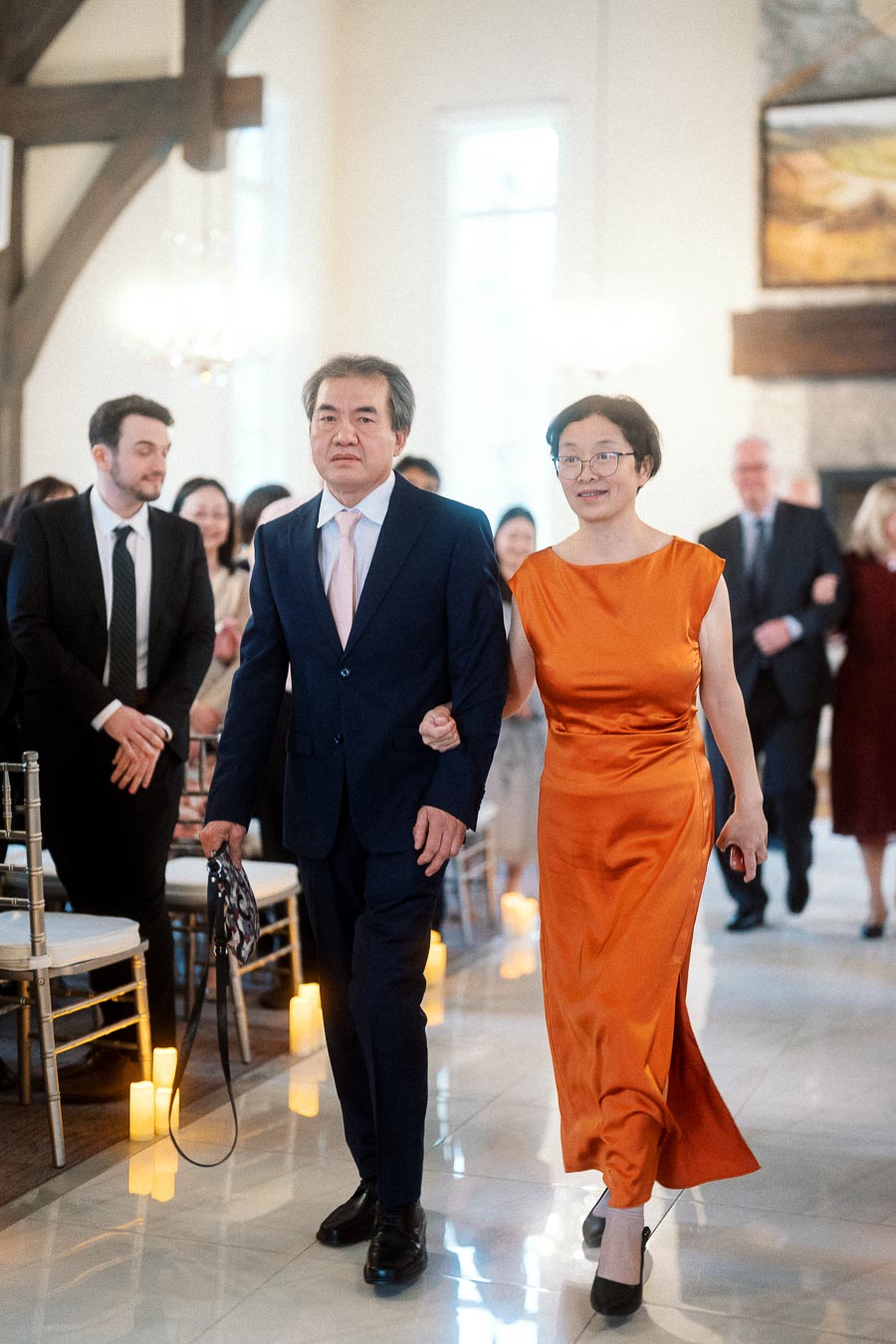 Two formally dressed people walk arm in arm down an indoor ceremony aisle.