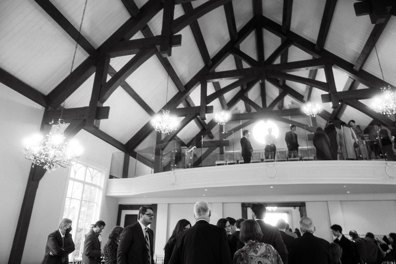 Guests gather beneath chandeliers and exposed beams in a high-ceilinged event hall.