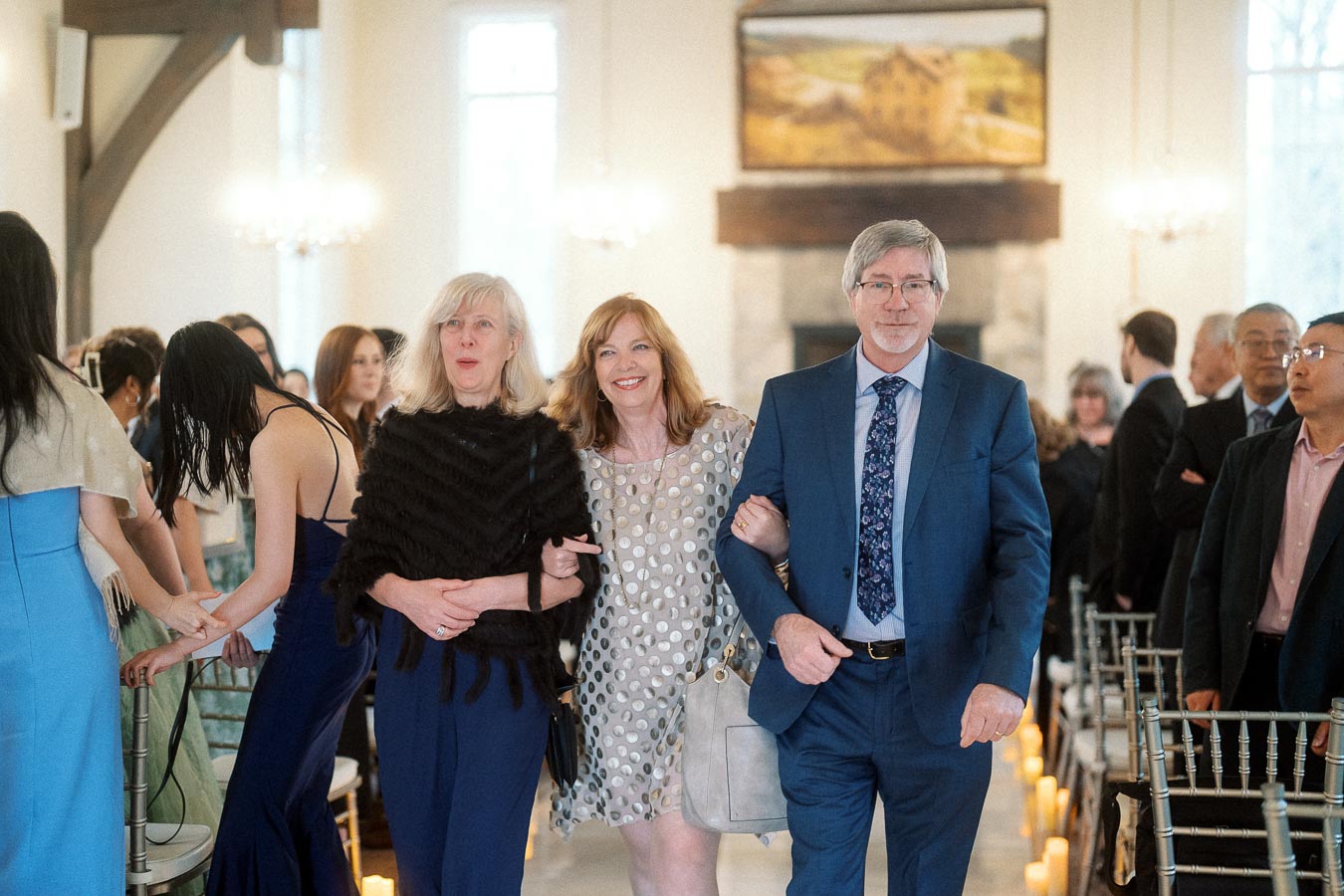 Three well-dressed guests walk arm in arm down an indoor ceremony aisle.
