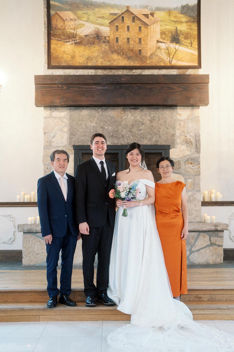 Couple in wedding attire posing with two family members in front of a stone fireplace